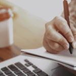 Image of a hand writing on a notepad with a laptop and coffee cup visible without the NEBOSH logo.